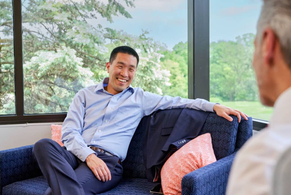 A man in a blue shirt smiles and sits on a blue couch, conversing with another person in an office with large windows and outdoor view.