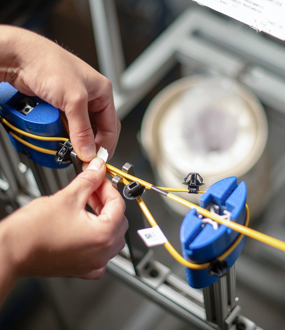 Hands applying labels to yellow wires secured in blue clamps within a mechanical setup.