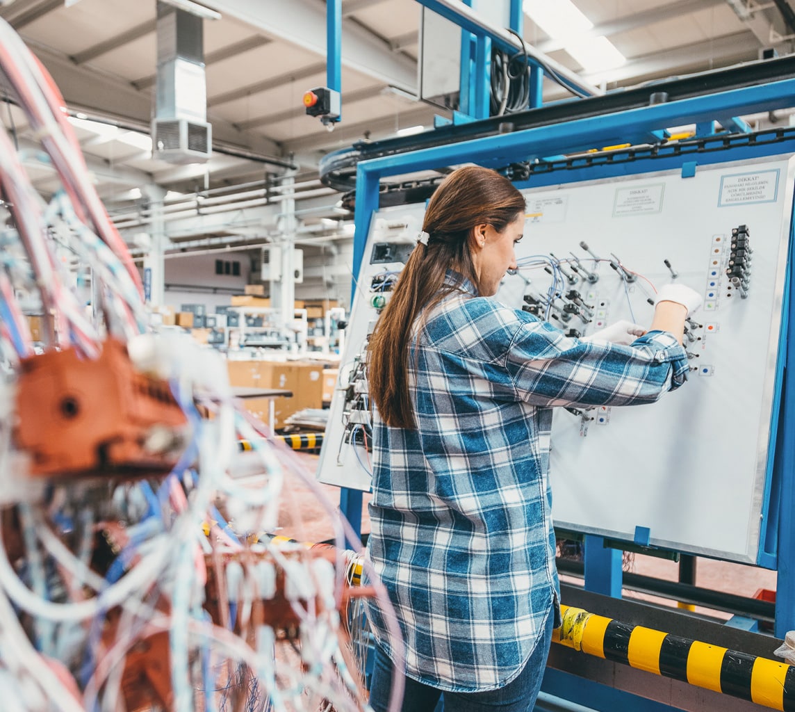 A woman in a plaid shirt works on wiring at a control panel in a modern industrial facility, surrounded by machinery and cables.