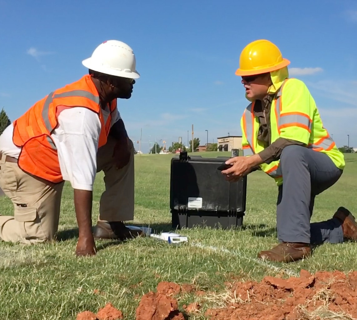 Two construction workers wearing hard hats and safety vests kneel on a grassy field beside a black equipment case, engaged in discussion.