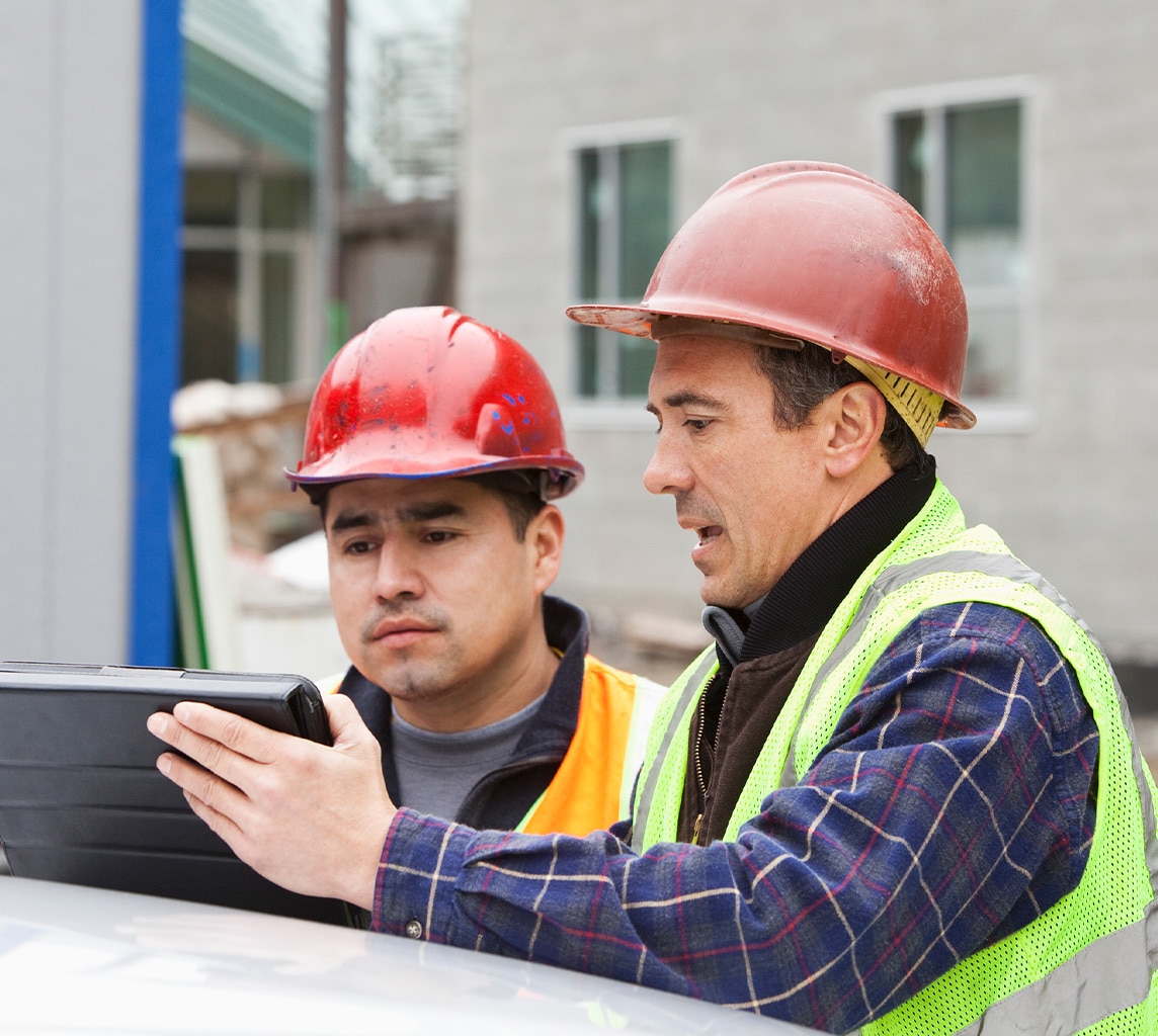 Two construction workers wearing hard hats and safety vests look at a tablet on a construction site.