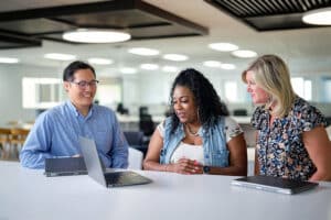 Three people are sitting at a table with open laptops, smiling and engaged in discussion, in a brightly lit office space.