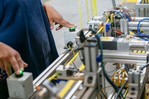 A person operates a green button on complex machinery with wires and components visible, likely in an industrial or manufacturing setting.