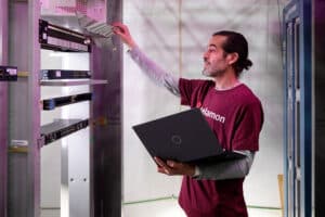 A person holding a laptop is inspecting a server rack, lifting a panel for a closer look. Shelves with electronic equipment are visible in the background.