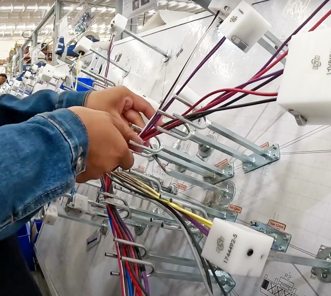 A person in a denim jacket is assembling and organizing electrical wires on a metal panel in an industrial setting.