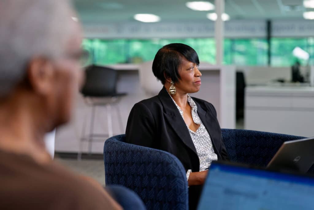 Two people are seated in an office setting, focusing on their laptops. The woman in the background is wearing a black blazer and earrings, while the person in the foreground is slightly out of focus.