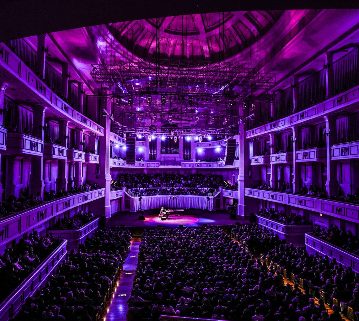 A large audience watches a performer on stage in an ornate theater lit with vivid purple lighting.