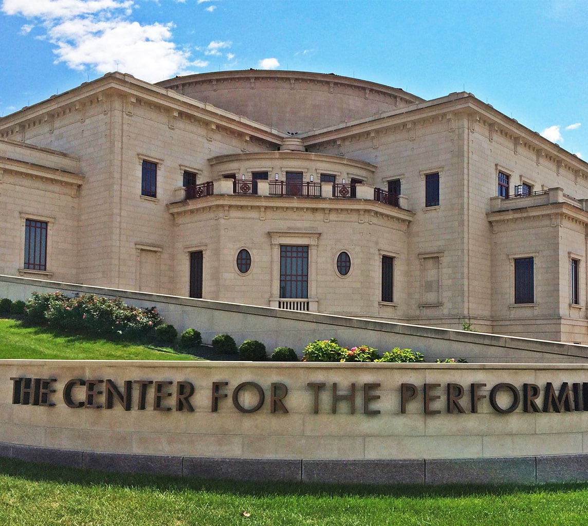 A stone building with columns titled "The Center for the Performing Arts," set against a blue sky with a manicured lawn and flowers in the front.