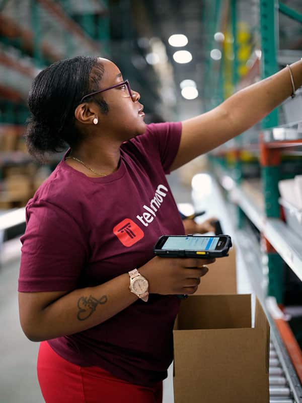 A person in a red Telmon shirt organizes inventory on shelves in a warehouse, holding a scanner in one hand and reaching for items with the other.