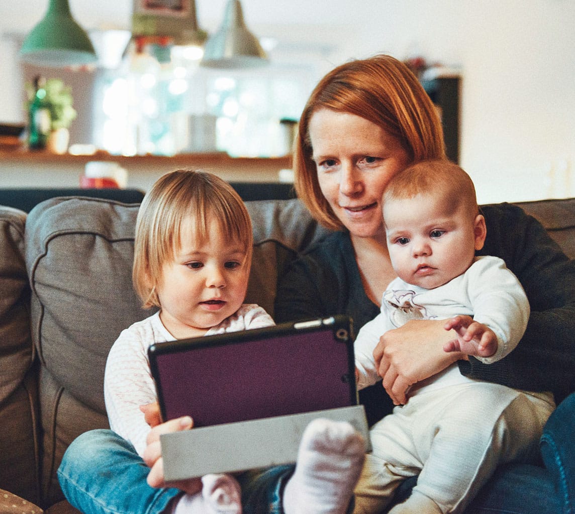 An adult holding a baby and a small child seated on a couch, all looking at a tablet screen together.