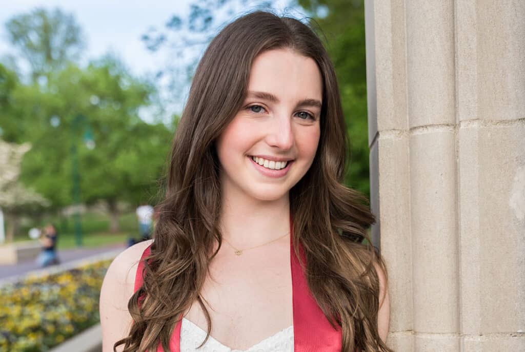 A young woman with long brown hair smiles at the camera while standing next to a stone pillar in an outdoor setting with trees in the background.