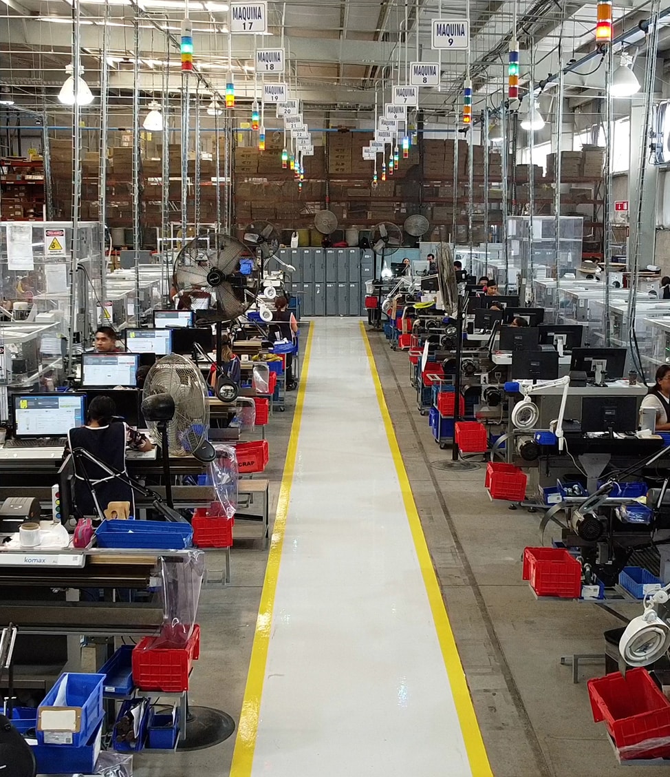 A factory floor with rows of workers operating machinery and computers. There is a central walkway with yellow lines, and overhead signs labeled "MAQUINA" identifying each station.