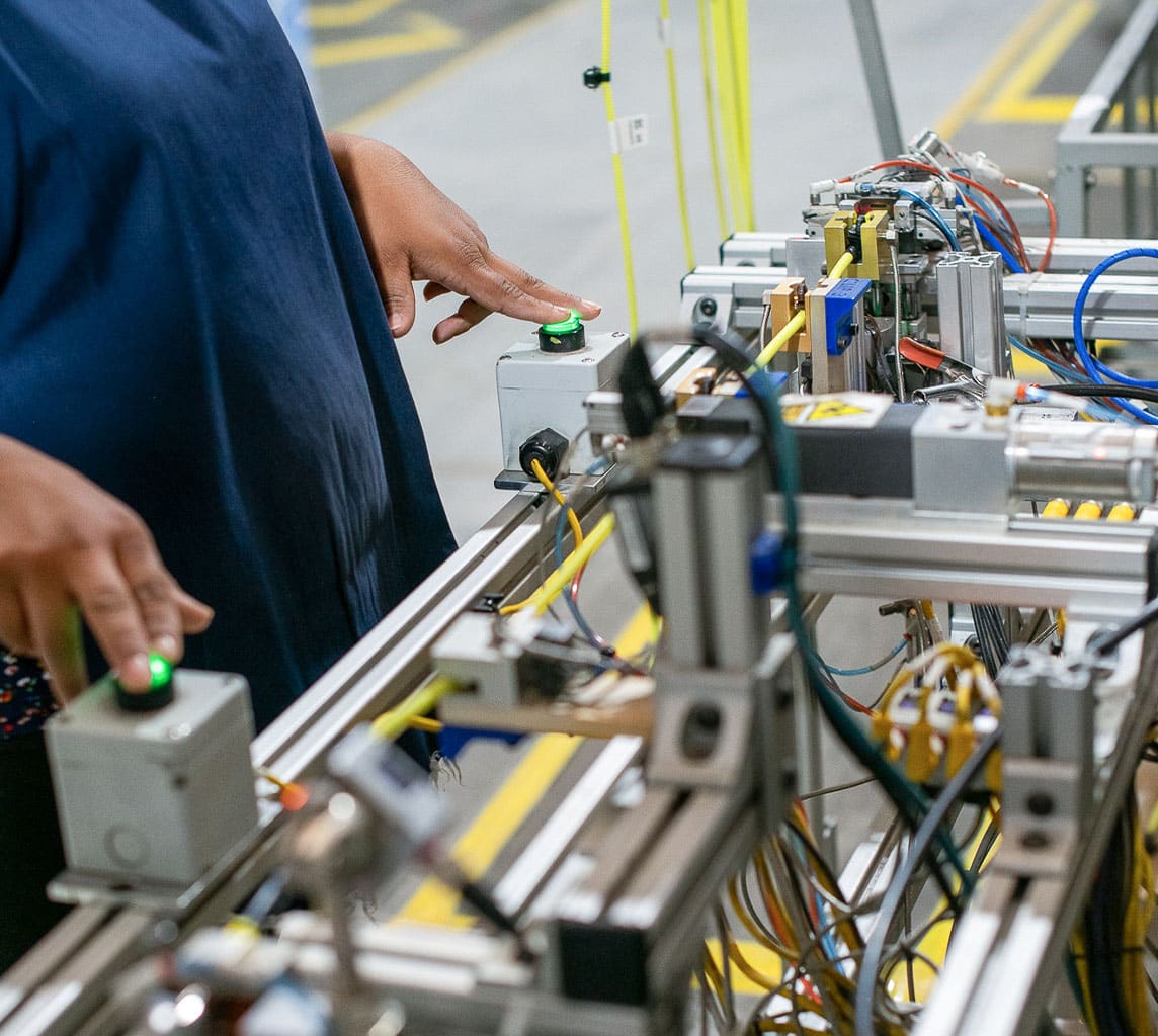 Person operating machinery with green button on an industrial production line, surrounded by cables and electronic components.