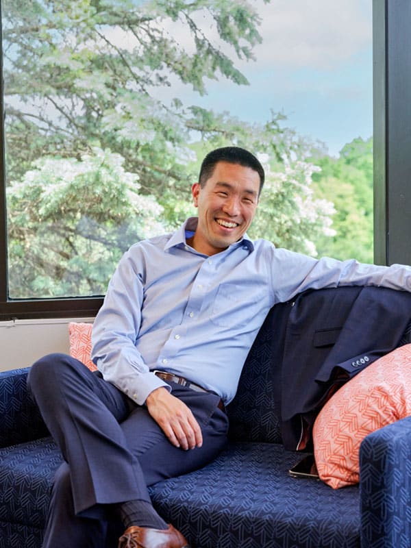 A man in business attire sits on a couch smiling with his arm resting on the back. Trees and sky are visible through the window behind him.