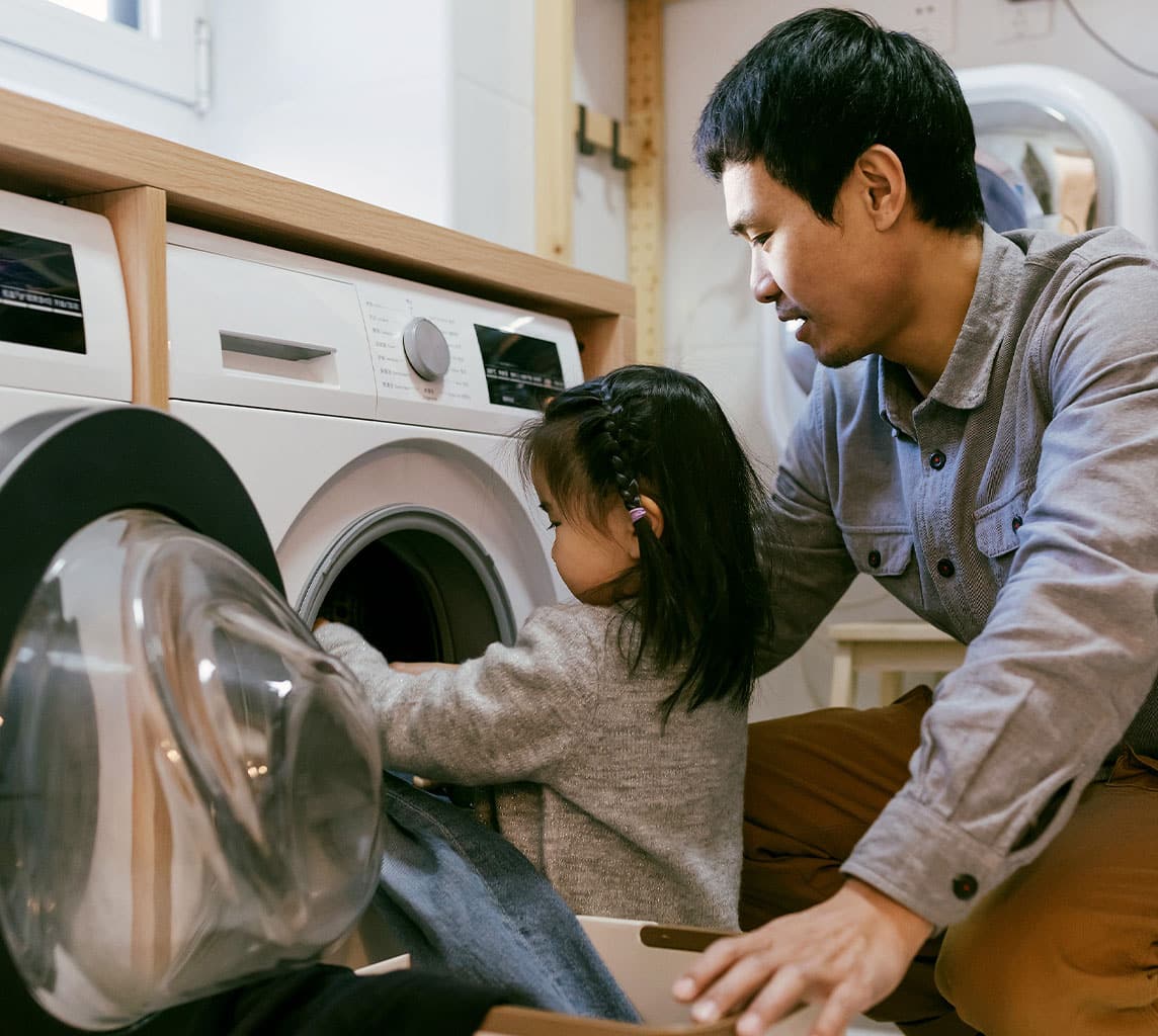 A man and a young child are loading clothes into a front-loading washing machine in a laundry room. The man kneels beside the child, who is placing a garment inside the washer.